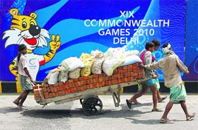 Labourers pull a hand cart loaded with bricks and sacks of sand in front of boards advertising the Games