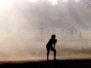 A young cricketer fields in the shade of a building as his teammates are engulfed in dust 