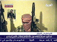 A masked man holds an automatic rifle in front of a wall which bears photographs of Saddam and his sons