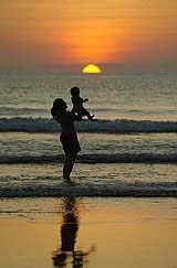 A woman lifts up her child as the sun sets on Kuta beach