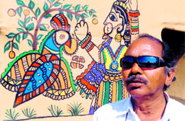  N K Sathi poses in front of a hut with Madhubani painted wall.