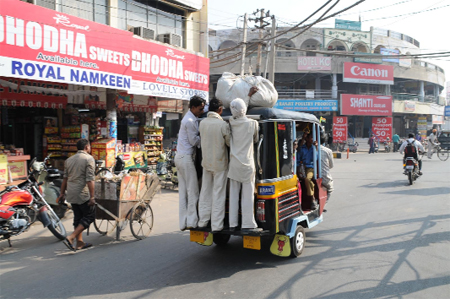 Three men travel perched on the footboard of an auto-rickshaw with their luggage resting atop the vehicle unmindful of the risks involved. 