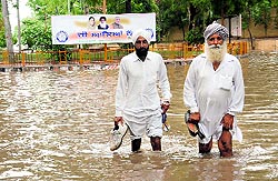 People wade through an inundated road with the backdrop of a poster carrying pictures of MP Harsimrat Kaur Badal, Chief Minister Parkash Singh Badal and Prime Minister Narendra Modi extending warm welcome to visitors near the DC and SSPs residences